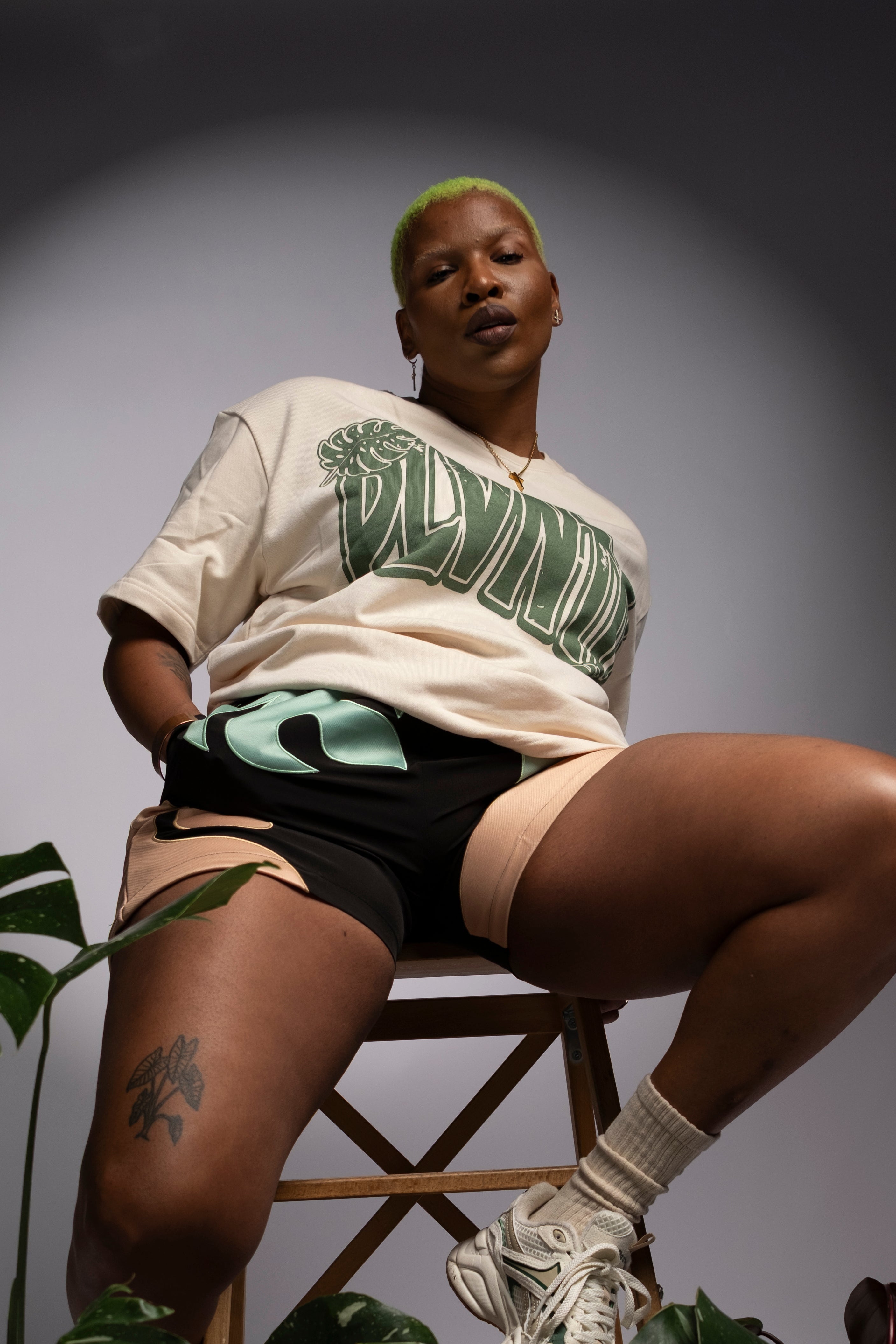 Person sitting on a stool with plants around, wearing a white t-shirt with green text and black shorts.